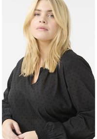 Blouse noire avec une coupe décontractée, tissu texturé avec de petits points en relief et des poignets élastiques. Décolleté en V et manches longues.