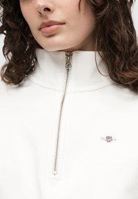 Femme aux cheveux bouclés portant un pull blanc à col haut avec une fermeture éclair partielle et un petit logo brodé rouge, blanc et bleu sur la poitrine.