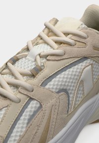 Μπεζ και λευκό αθλητικό παπούτσι με άνω μέρος από δίχτυ, σουέτ λεπτομέρειες και γκρι διακοσμητικά στοιχεία. Διαθέτει χοντρές κορδόνια και υφή πλαϊνών σχεδίων.