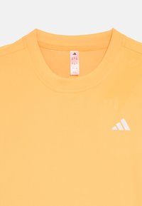 Jasnopomarańczowa sportowa koszulka z okrągłym dekoltem, krótkimi rękawami oraz białym logo Adidas na lewej piersi. Gładka tkanina z otworami wentylacyjnymi.