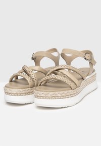 Sandali platform beige con cinturini incrociati, dettagli borchiati, una suola intermedia in juta intrecciata e un cinturino alla caviglia regolabile con fibbia in metallo.
