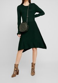 Mörkgrön långärmad stickad klänning med hög-låg fåll, i sällskap med en rund väska i ormskinn och beige, mönstrade ankelstövlar med klack.