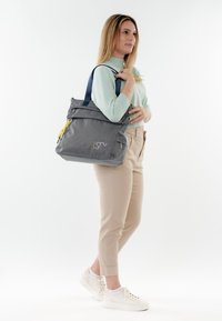 Donna in piedi che indossa pantaloni beige e un maglione verde chiaro, con una grande borsa tote grigia con manici blu navy e dettagli gialli.
