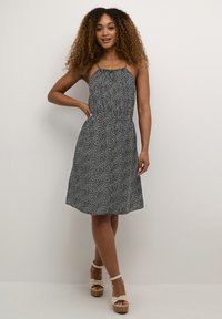 Robe à pois noir et blanc avec des bretelles spaghetti, taille cintrée et jupe évasée. Associée à des sandales à plateforme de couleur claire.