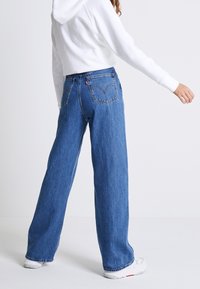 Blue denim jeans with a high-waisted, relaxed fit, featuring two back pockets and subtle stitching details. Worn with a white hoodie.