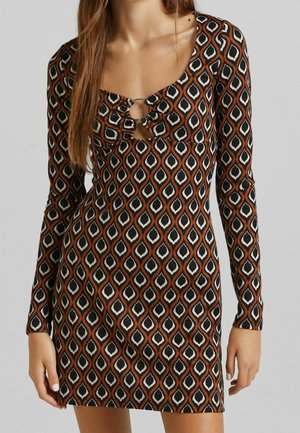 Woman wearing a long-sleeve mini dress with a brown, black, and white geometric pattern and a ring detail at the chest.