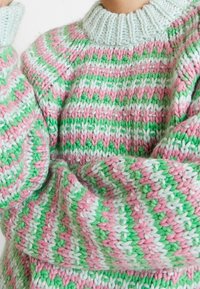 Suéter de punto en rayas rosas, verdes y blancas; cuenta con un cuello de canalé en azul claro; textura suave y gruesa; mangas largas y holgadas.