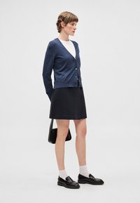 Navyblauwe cardigan met knopen aan de voorkant en geribbelde manchetten, gecombineerd met een zwarte A-lijn rok. Zwarte loafers en een kleine zwarte tas maken de outfit compleet.