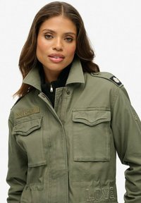 Chaqueta militar de color verde oliva, con cierre de cremallera, dos bolsillos en el pecho, parches en los hombros y detalle bordado de "LOVE" en la parte inferior.