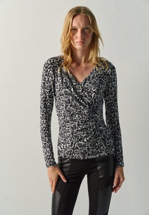 Mujer rubia que lleva una camiseta de manga larga con estampado en blanco y negro y pantalones negros con detalles de parches de cuero texturizado, de pie contra una pared lisa.