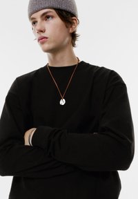 Schwarzer Sweatshirt mit langen Ärmeln und rundem Ausschnitt, kombiniert mit einer braunen Lederkette, die einen metallischen ovalen Anhänger trägt.