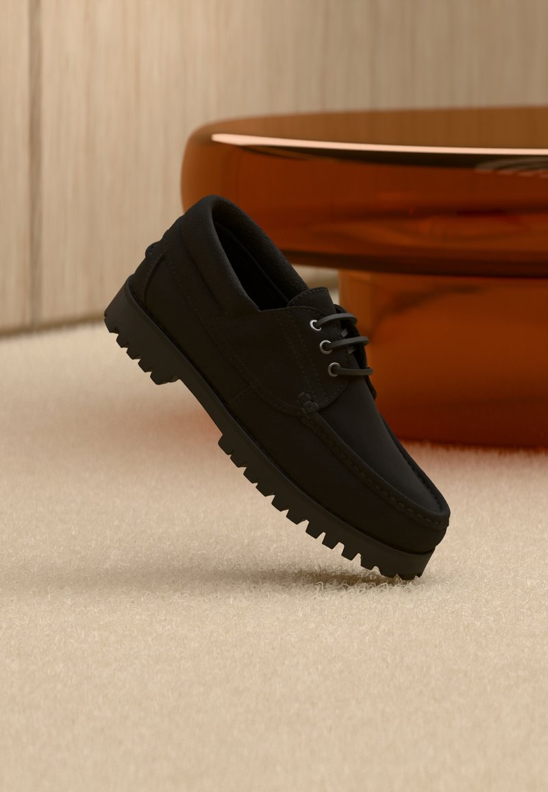 Chaussure noire à lacets avec semelle épaisse à nervures flottant au-dessus d'un tapis beige, table ronde moderne marron en arrière-plan dans une pièce à éclairage doux.