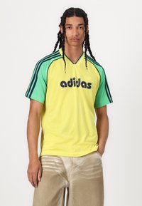 Svetlo rumena mrežasta majica z zelenimi rokavi, s črnim logotipom "adidas" spredaj. Kratki rokavi, oblikovanje z V-izrezom in zračen material.