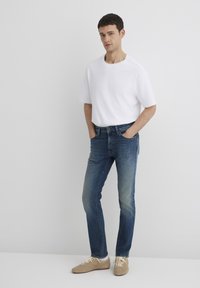 T-shirt bianca a maniche corte, jeans in denim blu con lavaggio sbiadito e scarpe color beige chiaro. Il modello è in piedi contro uno sfondo bianco semplice.