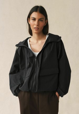 Frau mit dunklen Haaren, die eine schwarze Kapuzenjacke mit Reißverschluss über einem weißen Oberteil und braunen Hosen trägt, vor einem einfarbigen beigen Hintergrund stehend.