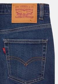 Vaqueros de mezclilla azul oscuro con costuras naranjas, con un parche de cuero etiquetado "Levi Strauss & Co." y una pestaña roja en el bolsillo trasero.