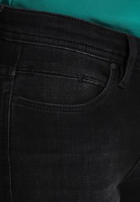 Jeans de mezclilla negra con un diseño ajustado, que cuentan con un pequeño bolsillo y detalles de costura. El material parece ligeramente texturizado y duradero.