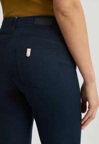 Traperice tamnoplavih nijansi imaju elegantan dizajn s vidljivim stražnjim džepom ukrašenim malim metalnim oznakom brenda. Glatka tekstura, prilagođen oblik.