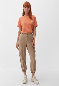 Camiseta de algodón naranja con un pequeño logo, pantalones cargo beige con puños elásticos y bolsillos laterales, combinados con zapatillas gruesas beige.