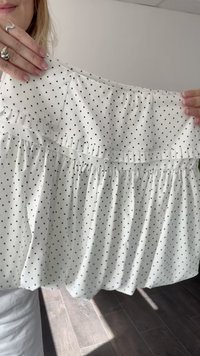White blouse with black polka dots, featuring ruffled design, gathered waist, and soft fabric texture, held in hands against a light background.