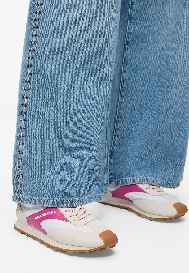 Baskets avec une tige en maille blanche, des accents roses, un capuchon en beige et une semelle texturée brune. Associées à un jean bleu clair avec des détails en clou.