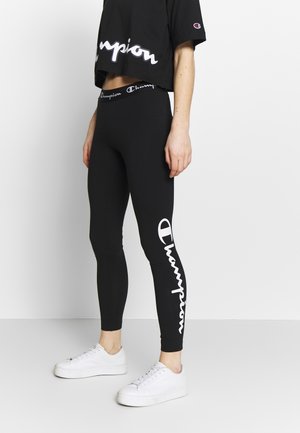 Persona con leggings negros de Champion y top corto con texto del logo en blanco, combinados con zapatillas de plataforma blancas, de pie frente a una pared lisa.