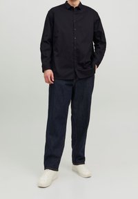Camicia nera con colletto classico, abbinata a jeans scuri a vestibilità ampia e sneakers bianche. Tessuto liscio.