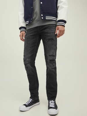 Jeans de mezclilla negros con un corte ajustado, que presentan detalles desgastados y desgarros visibles. Combinados con zapatillas de lona negras y una chaqueta tipo varsity azul marino.