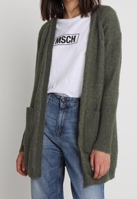 Cardigan lavorato a maglia verde oliva con apertura frontale, polsini a coste e due tasche frontali, indossato sopra una maglietta bianca e jeans blu.