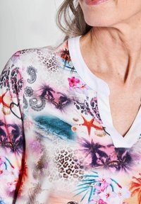 Korte mouwen top met een kleurrijke bloemen- en strandprint en een witte V-hals kraag. Materiaal lijkt zacht met een gladde textuur.