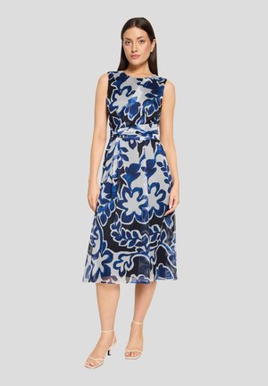 Vrouw die een mouwloze, knielange jurk draagt met een blauw-zwart abstract bloemmotief, gecombineerd met beige enkellaarsjes met bandje bij de enkel, staande op een grijze achtergrond.