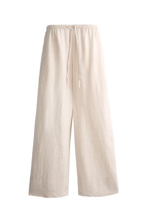 Femme debout portant un haut court blanc sans manches, un pantalon beige ample avec cordon de serrage, et des sandales noires sur un sol en pierre claire.