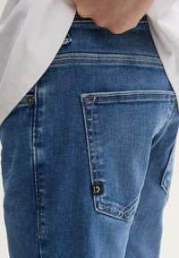 Vaqueros de mezclilla en azul oscuro con un corte slim. Las características incluyen cinco bolsillos, costuras en contraste y una pequeña etiqueta negra en el bolsillo trasero.