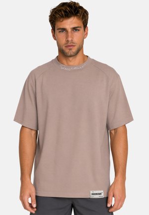 Mann mit kurzem welligem Haar trägt ein lockeres beiges T-Shirt mit dem Label "Reichstadt" nahe am Saum und passendem Text am Kragen, der vor einem einfarbigen Hintergrund steht.