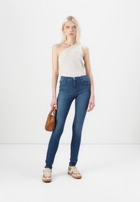Top beige de un solo hombro, jeans de denim ajustados, de cintura alta, azul oscuro, con textura suave, combinados con zapatillas beige y un bolso pequeño marrón.