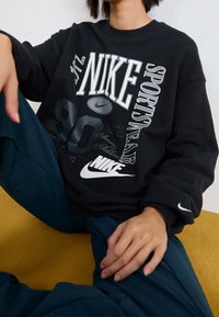 Person sitzt mit einem gebeugten Bein, trägt einen schwarzen Nike-Hoodie mit weißem und grauem sportlichem Text und Logos sowie dunkelblauen Hosen.