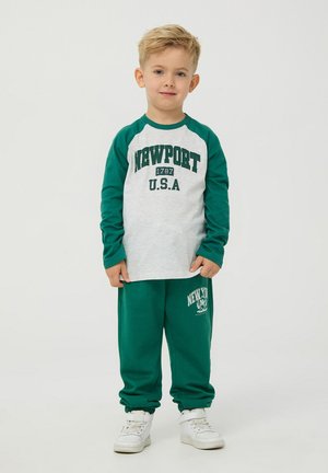 Maglietta raglan verde e grigia con stampa "NEWPORT U.S.A.", abbinata a pantaloni sportivi verdi con logo decorativo. Sneakers bianche.