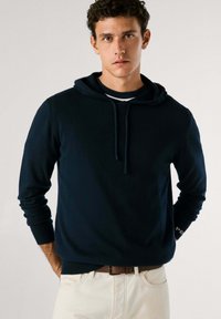 Temno modra kapuca iz mehkega materiala z sprednjim kenguru žepom, rebrastimi manšeti in vrvico. Oblačena čez črtasto srajco.