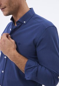 Camisa azul marino de botones, hecha de una tela texturizada, con cuello, botones blancos y un logo bordado en el pecho.