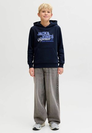 Garçon blond debout portant un sweat à capuche bleu marine "Jack & Jones", un jean large gris et des baskets blanches sur un fond clair uni.