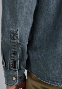 Koszula dżinsowa w wyblakłym niebieskim kolorze, z guzikami na mankietach, kontrastowym szwem i metką na rękawie. Miękka struktura i luźny krój.