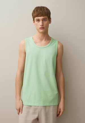 Tânăr cu păr scurt și castaniu, purtând un tricou verde deschis fără mâneci și pantaloni bej, în picioare în fața unui fundal simplu.