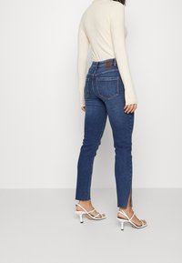 Höga blå denimjeans med sidoflikar; kombinerade med en beige långärmad topp och vita sandaler med remmar.