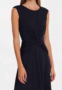 Mörkblå ärmlös klänning med rundad halsringning och snurrat tygdetalj vid midjan, tillverkad av mjukt, töjbart material.