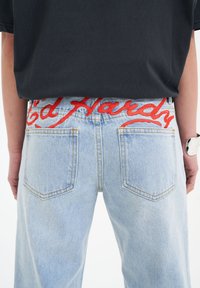 Jeans in denim azzurro chiaro con un logo rosso "Ed Hardy" sul girovita. Presentano tasche posteriori standard e dettagli di cucitura visibili.
