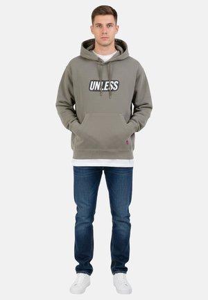 Giovane uomo in piedi che indossa una felpa con cappuccio grigia con la scritta "UNLESS", jeans blu, sneakers bianche e una maglietta bianca sotto, su sfondo semplice.