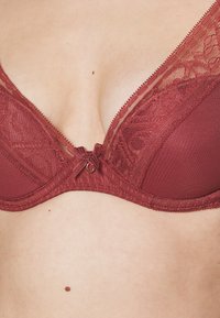 Sutiã de renda burgundy e tecido com detalhes transparentes, copas triangulares e um laço central. Apresenta renda texturizada e acabamento.