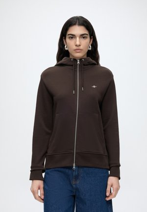 Kvinde med langt mørkt hår iført sølvfarvede øreringe i ringe, en mørkebrun hættetrøje med lynlås og blå jeans mod en ensfarvet hvid baggrund.