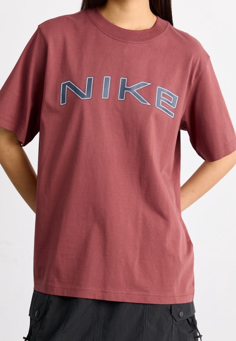 Person som bär en lös maroon Nike t-shirt med djärva navy och vita broderade bokstäver över bröstet, tillsammans med svarta cargobyxor.