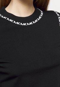 Haut en tricot noir avec motif répété du logo "MK" en blanc le long de l'encolure ronde et de la couture de l'épaule, porté par une personne aux cheveux longs.